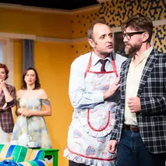 Na scenie "Mayday och teatr" mężczyzna w fartuszku z gęśmi namawia kolegę w kratę. W tle panie.