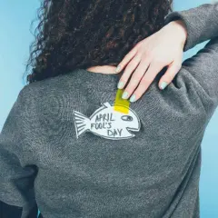 Ręka z pastelowym lakierem przyczepia do swetra papierową rybę z napisem "April Fool's Day".