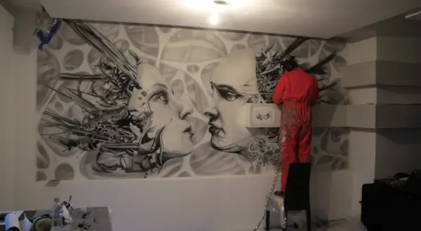 Artysta w czerwonym kombinezonie tworzy malarstwo artystyczne wnętrz – surrealistyczny mural z twarzami i mechanicznymi elementami.