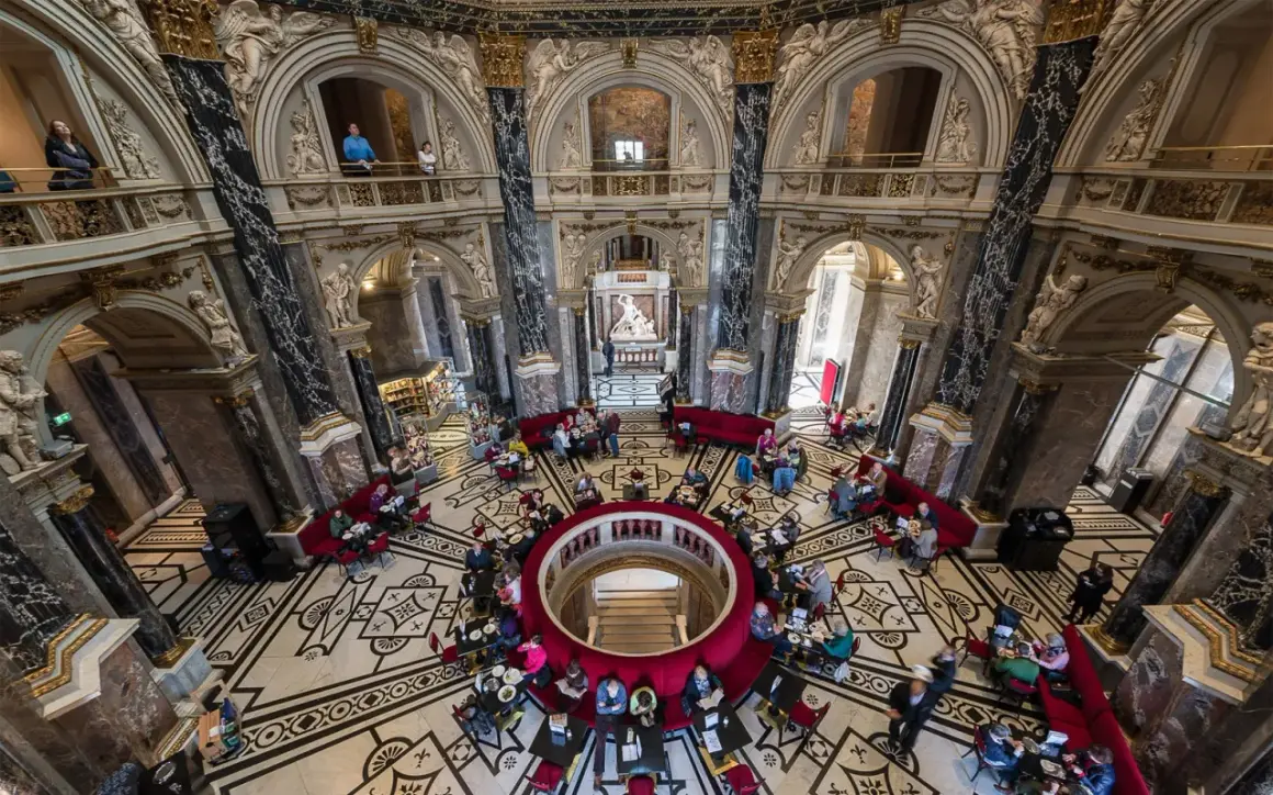 Wnętrze muzeum historii sztuki w Wiedniu z ludźmi przy stolikach i rzeźbą w centrum.