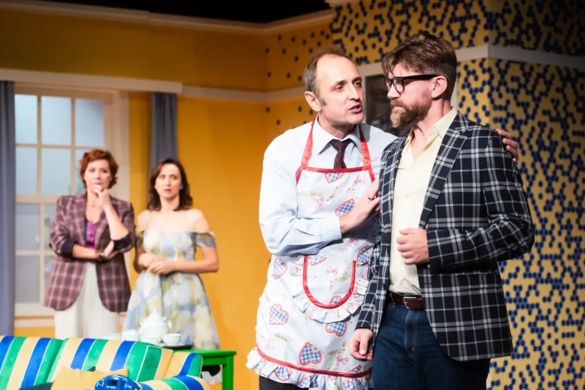 Na scenie "Mayday och teatr" mężczyzna w fartuszku z gęśmi namawia kolegę w kratę. W tle panie.
