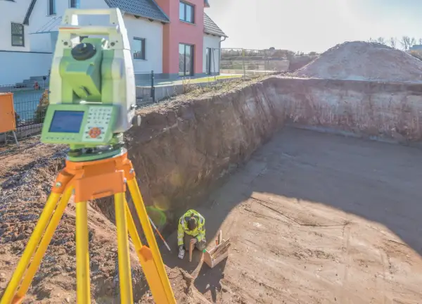 Geodeta dokonuje pomiaru rzeźby terenu na budowie. Widać niwelator i pracownika w wykopie.