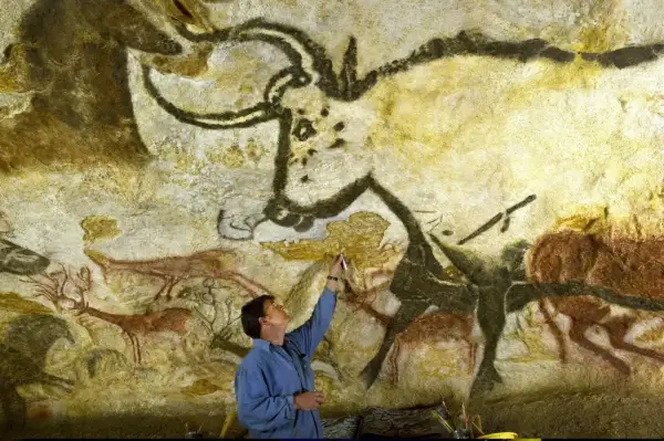 Człowiek bada prehistoryczne malowidła z Lascaux, dotykając pędzlem skalnej ściany z wizerunkami zwierząt.